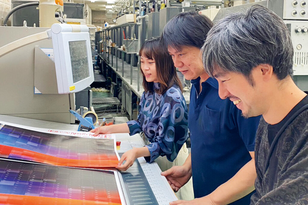 写真右より橋詰冬樹氏（グラフィックデザイナー）・谷井印刷機機長・西谷内PD。10色印刷機オペレーションテーブルにて印刷立ち合い、色調確認の様子。